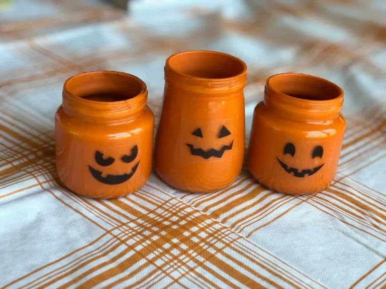 Baby food jars painted orange with Jack-O-Lantern faces on them sitting on a white and orange plaid tablecloth