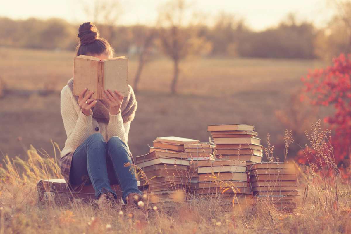 A woman sits with a stack of books in a field and is reading