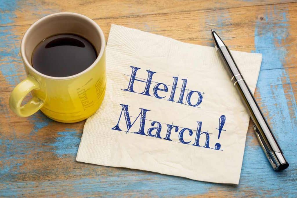 Cup of coffee in a yellow cup on a table with a napkin and pen and the napkin reads "Hello March"