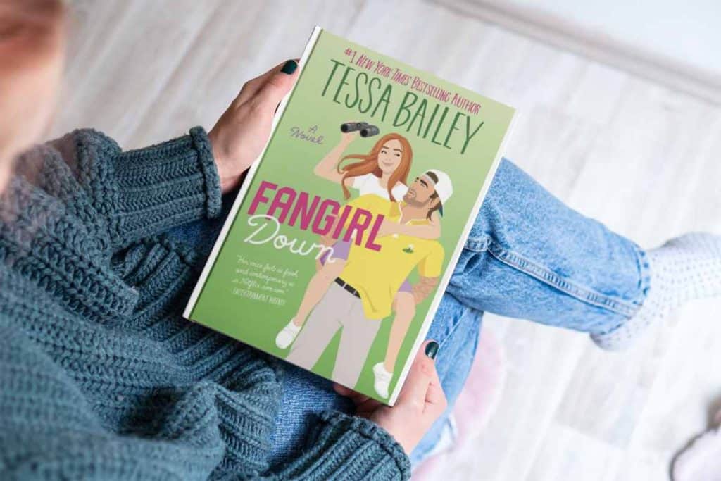 A woman sitting with her legs crossed with a book in her lap called "Fangirl Down"