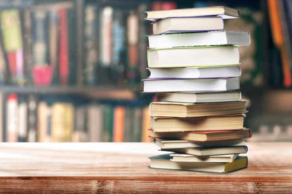 Stack of books on a wooden table in front of a bookshelf stuffed full of books.
