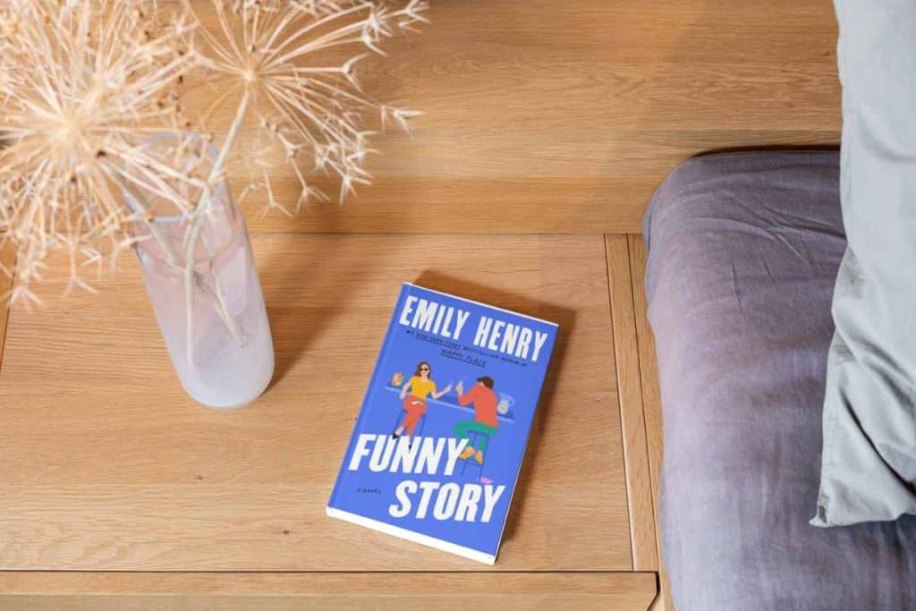 A book titled "Funny Story" on a bedside table with a vase next to it