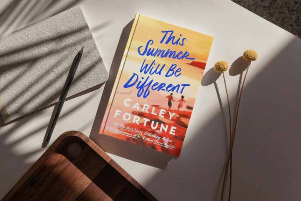 Book titled "This Summer Will Be Different" sitting on a table with a paper and pen and wooden tray and a flower
