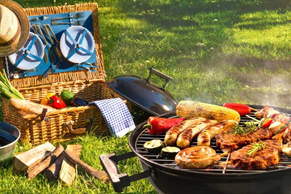 A barbeque grill cooking all kinds of meats and vegetables with a picnic basket open in the background.
