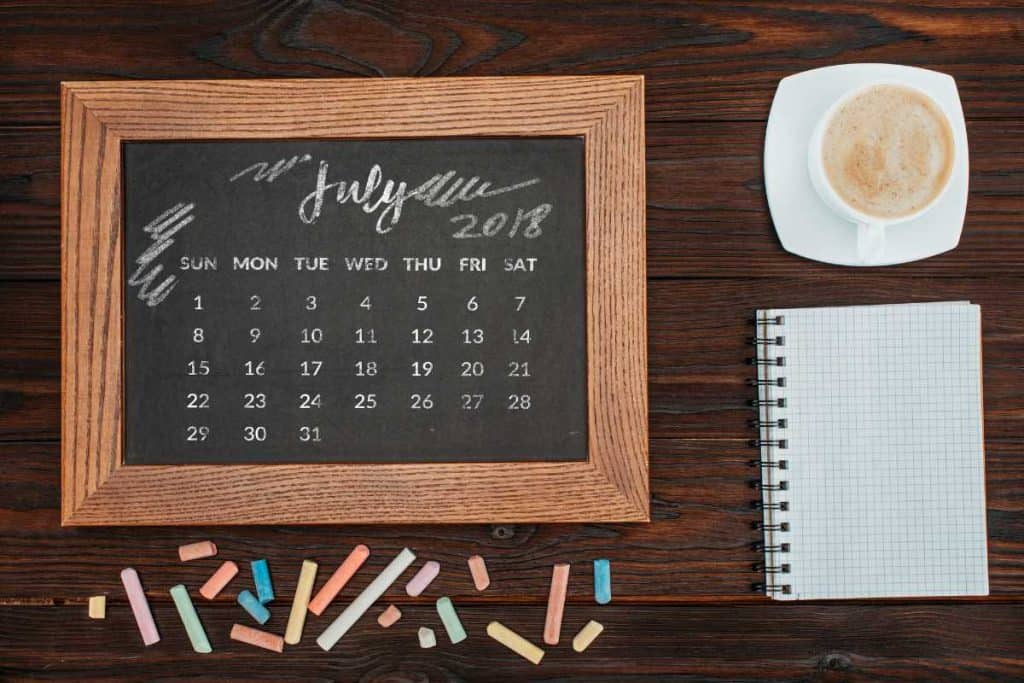 Chalkboard calendar with the month of July written on it along with a notebook and coffee on a wooden table, with some colored chalk on the side.