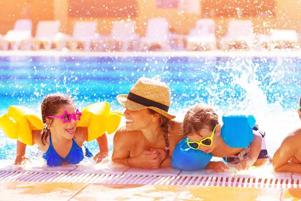 A family gathered in the pool by the side of the pool splashing and having fun. The kids are wearing sunglasses.