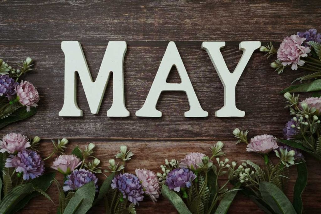 Block letters spelling MAY with purple flowers lying on a wooden table