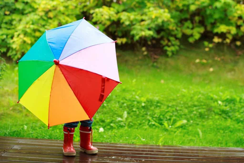 Little kid wearing red rain boots holidng a rainbow colored umbrella in front of them