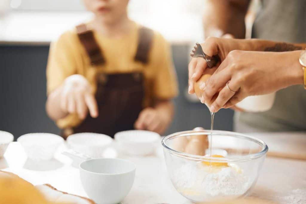 Woman cracking an egg into a bowl with flour and a kid in the background watching