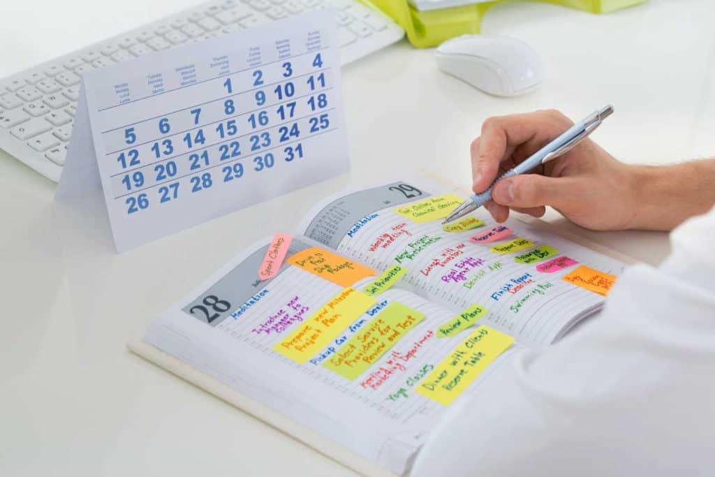 Calendar and agenda book open on a desk with lots of colorful notes nad post it notes on the page witha white computer keyboard and mouse in the background.
