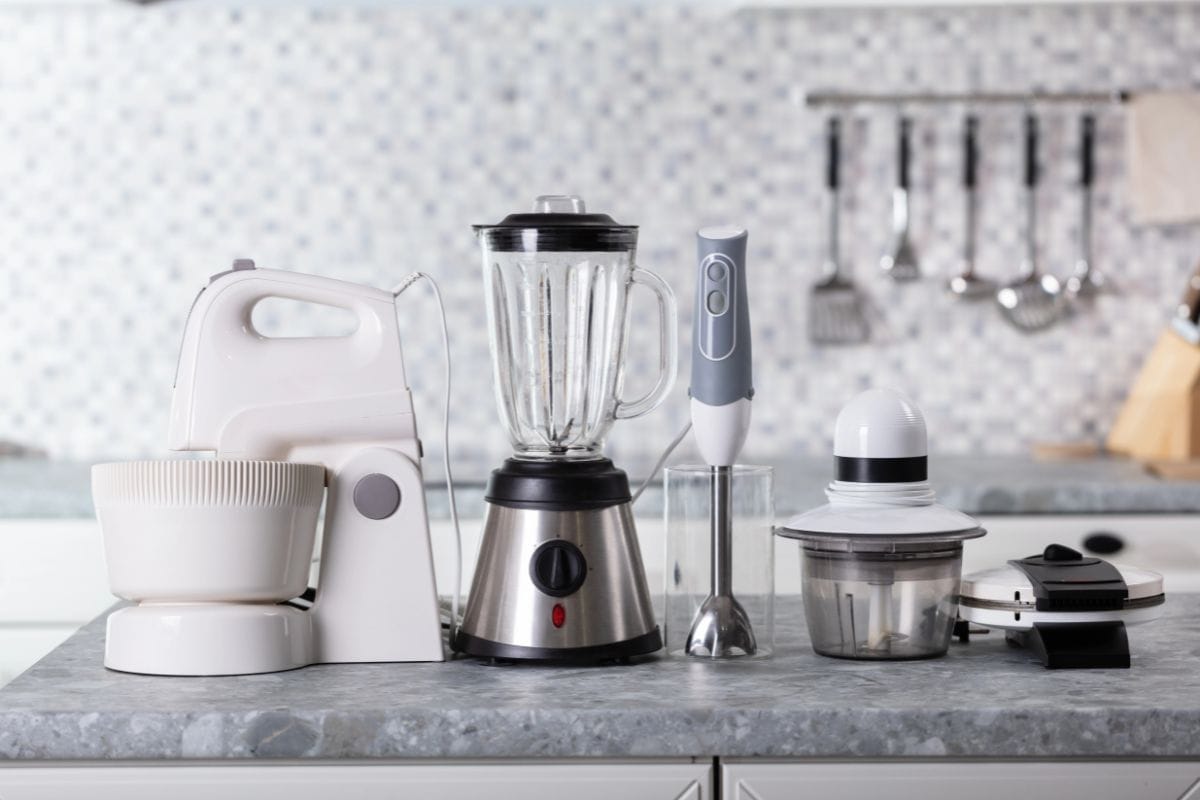 Small appliances sitting on the gray countertop including: a stand mixer, a blender, an immersion blender, a food processor and a waffle maker.