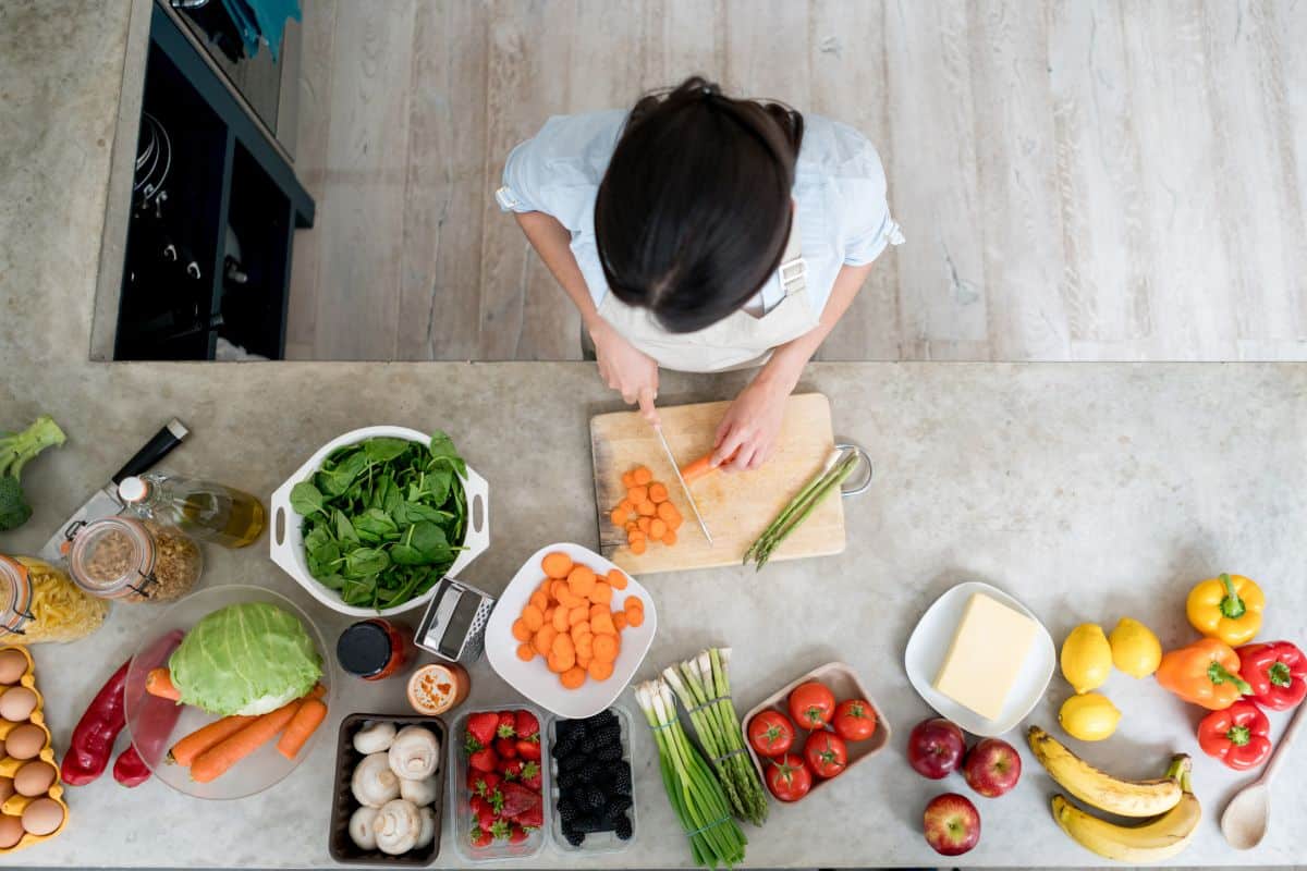 Shot from above, a woman is cutting and preeping a salad with a knife and cutting board and is surrounded by all kinds of fruit and vegetables.