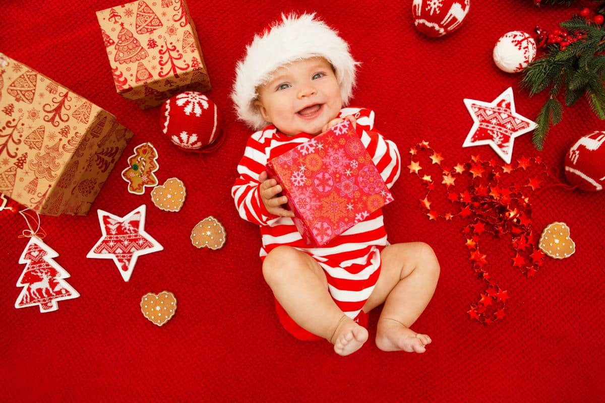 Smiling baby with a santa hat laying on a red blanket surrounded by Christmas items like cookies and ornaments and baby his holding a wrapped present and smiling