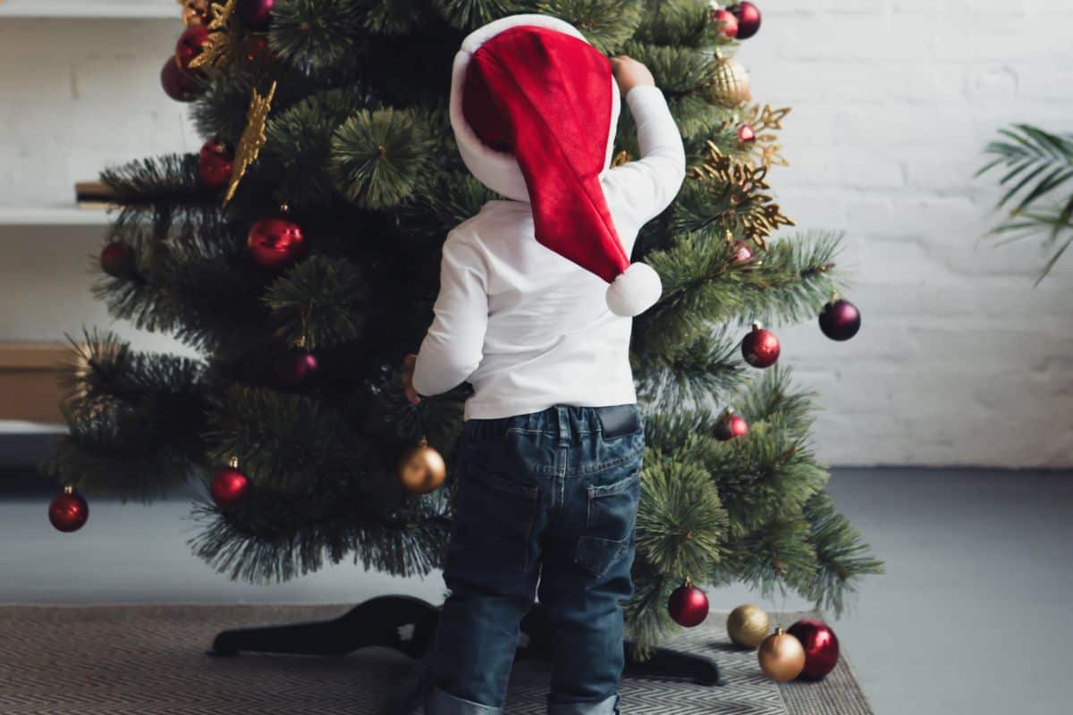 Toddler standing in front of the Christmas tree putting on an ornament wearing a santa hat and white shirt and jeans