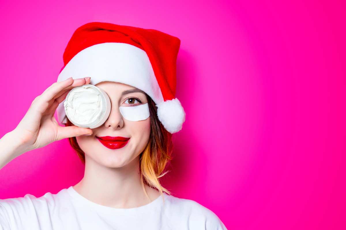 A woman with beauty eye patch on one eye and a container of face cream on the other eye, wearing a Santa hat