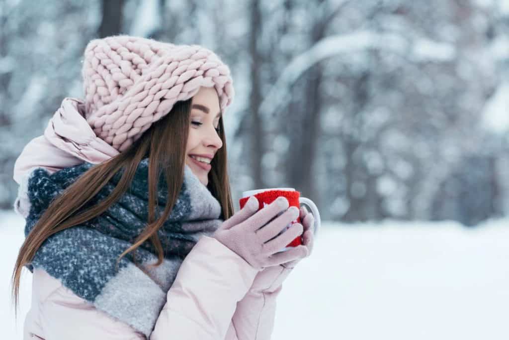 A woman dressed in winter jacket, hat and coat sipping a mug of hot chocolate.